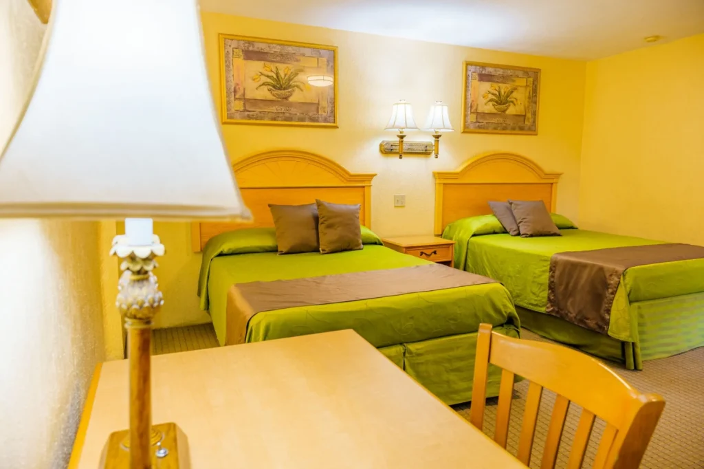 Hotel room with two double beds featuring green bedding and brown throws, wooden desk with lamp, chair, and framed botanical paintings on the wall.