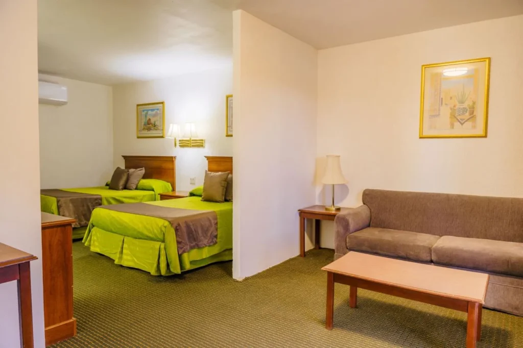 Hotel suite room with two double beds in green and brown bedding, a brown sofa, wooden coffee and side tables, a lamp, and framed wall artwork.