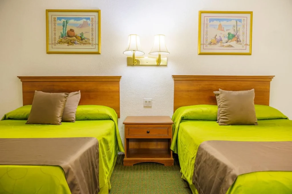 Hotel room with two twin beds with green covers and beige pillows, a wooden nightstand with a drawer, a double wall lamp, and framed artwork above each bed.