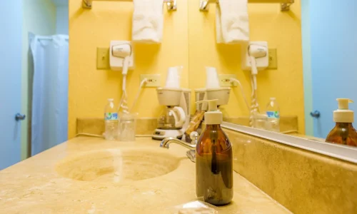 Hotel bathroom countertop with sink, soap dispenser, coffee maker, bottled water, glasses, hairdryer, and towels reflected in a mirror.