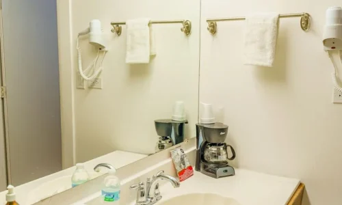 Hotel bathroom countertop with a sink, soap dispenser, water bottle, disposable cups, coffee maker with cups, wall-mounted hairdryer, and towels on a rack.