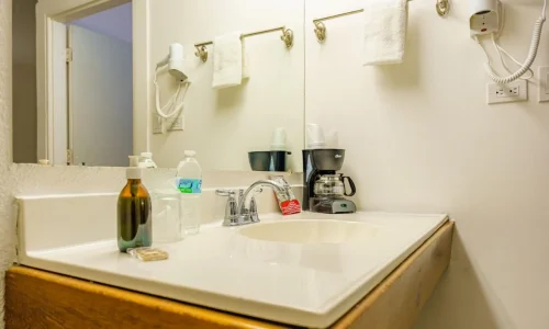 Hotel bathroom sink area with mirror, hair dryer, towel rack with towel, soap dispenser, water bottle, and coffee maker with cups.