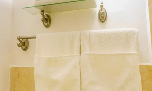 White towels hanging on a metal towel bar and folded on a glass shelf below a wall-mounted light in a bathroom with tiled walls.