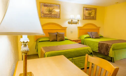 Hotel room with two double beds featuring green bedding and brown throws, wooden desk with lamp, chair, and framed botanical paintings on the wall.