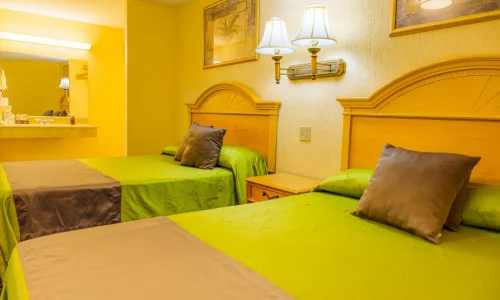 Hotel guest room with two beds featuring green bedspreads and brown pillows, wooden headboards, wall lamps, and a sink area with mirror.
