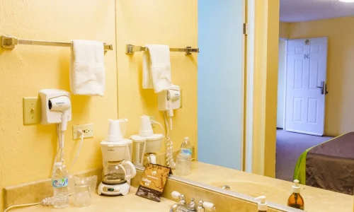 Hotel bathroom vanity with sink, countertop, large mirror, mounted hairdryer, towel, coffee maker with cups, bottled water, and soap dispenser, with view into hotel room.