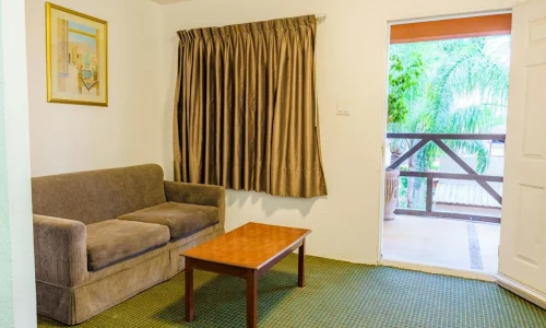 Hotel living room with brown sofa, wooden coffee table, closed brown curtains, wall painting, and open door leading to balcony with palm trees visible outside.