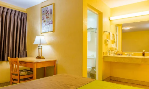 Hotel room featuring a bed, wooden desk with lamp and chair, dark curtains, and a bathroom area with sink, mirror, and toilet.