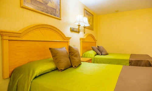 Hotel room with two double beds with wooden headboards, green bedspreads, brown pillows, a wooden nightstand, wall-mounted lamp, and framed artwork.