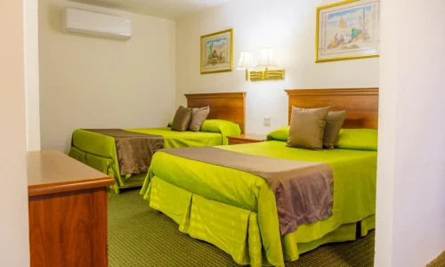 Hotel room with two beds dressed in green bedding and brown runners, wooden headboards, bedside table with lamps, framed artwork on white walls, and a wooden dresser.