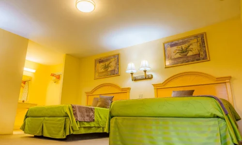Hotel room with two double beds dressed in green bedspreads, yellow walls with floral artwork, and a lighted sink area in the background.