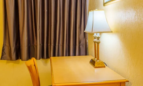 Wooden desk and chair with a lamp, in a hotel room with closed brown curtains and floral artwork on a yellow wall.
