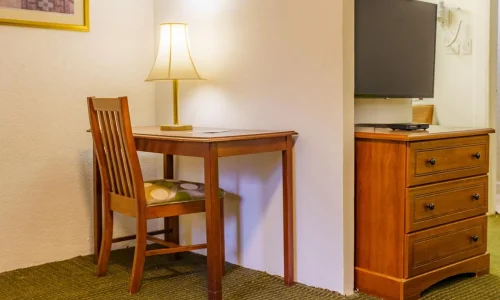 Hotel room corner with wooden desk, chair, table lamp, dresser, and wall-mounted flat-screen TV.