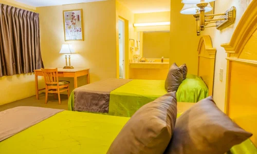 Hotel room with two beds featuring green and brown bedding, a wooden desk with chair and lamp, yellow-toned walls, and a vanity area with mirror and sink.