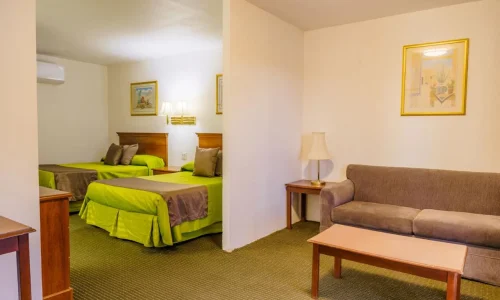 Hotel suite room with two double beds in green and brown bedding, a brown sofa, wooden coffee and side tables, a lamp, and framed wall artwork.