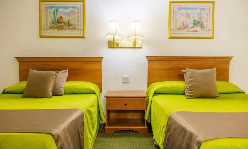Hotel room with two twin beds with green covers and beige pillows, a wooden nightstand with a drawer, a double wall lamp, and framed artwork above each bed.