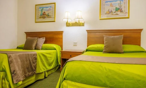 Hotel room with two beds, wooden headboards, lime-green bedding, taupe pillows and bed runners, wooden nightstand, double wall lamps, and two framed desert-themed paintings above beds.