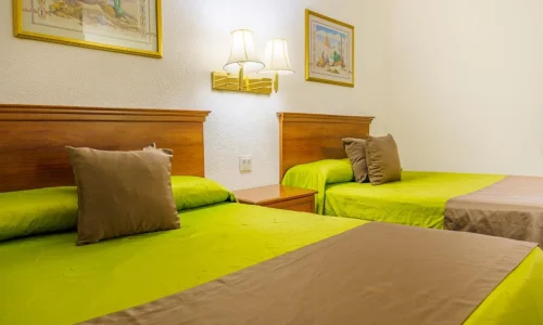 Two hotel beds with green bedding and beige throws in a room featuring wooden headboards, pillows, wall lamps, and framed artwork.